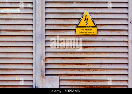 Two yellow triangular high voltage warning signs are mounted on a weathered gray metal louvered industrial door. Each sign displays a black lightning Stock Photo