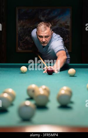 Concentrated mature man playing billiards, carefully aiming his cue stick to meet the white ball on a green pool table in a dimly lit space, showcasin Stock Photo