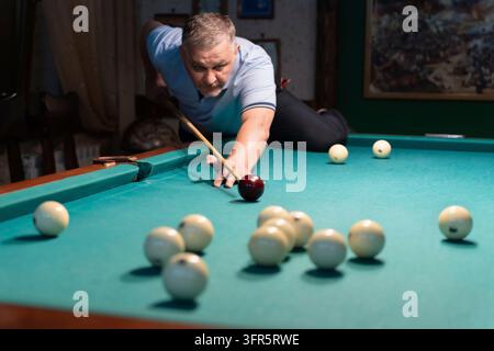 Concentrated mature man playing billiards, carefully aiming his cue stick to meet the white ball on a green pool table in a dimly lit space, showcasin Stock Photo