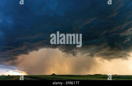 Intense rain falls from a dark supercell storm cloud over a lonely house in the plains, with the setting sun illuminating the precipitation from behin Stock Photo