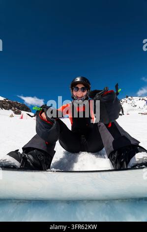 Cheerful female snowboarder laughing in winter mountains. Girl sitting and smiling on ski slope, preparing to jump Stock Photo