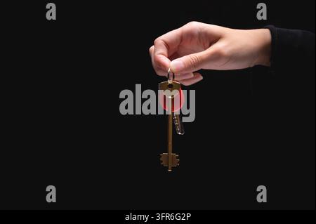 A hand holds the keys to the apartment on a black background. Young adult woman holding metal door keys in her hand, close-up. Blank space for text Stock Photo