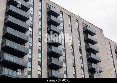 Low angle view of a modern brick rise high with stacked dark balconies and tall windows rising against a bright cloudy sky Stock Photo