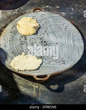 Top view of two handmade corn tortillas cooking on a traditional comal griddle in Ek Balam, Yucatan, Mexico. The uncooked masa rounds rest on a worn m Stock Photo