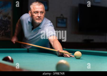 Concentrated mature man playing billiards, carefully aiming his cue stick to meet the white ball on a green pool table in a dimly lit space, showcasin Stock Photo