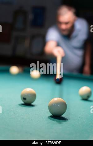Concentrated mature man playing billiards, carefully aiming his cue stick to meet the white ball on a green pool table in a dimly lit space, showcasin Stock Photo
