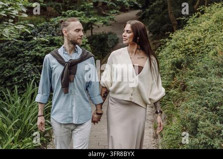 Outdoor shot of young couple in love walking on pathway through grass field. Young couple strolling in the park. Trendy couple with tattoos walking in Stock Photo