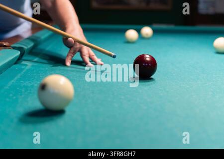 Concentrated mature man playing billiards, carefully aiming his cue stick to meet the white ball on a green pool table in a dimly lit space, showcasin Stock Photo