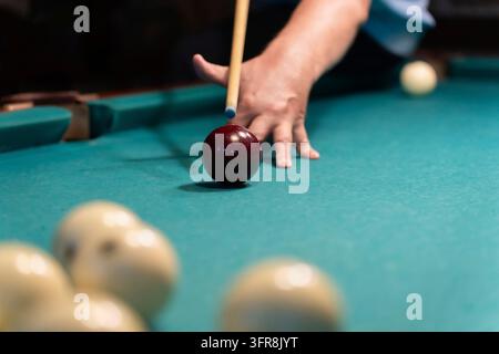 Concentrated mature man playing billiards, carefully aiming his cue stick to meet the white ball on a green pool table in a dimly lit space, showcasin Stock Photo