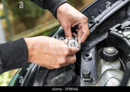 A person carefully handles a halogen headlight bulb with both hands near an open car hood during headlight maintenance work. The close-up perspective Stock Photo