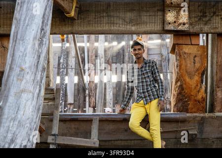 A young shipwright wearing yellow trousers sits on a wooden beam inside a boatyard smiling for the camera, Gujarat, Mandvi, India Stock Photo