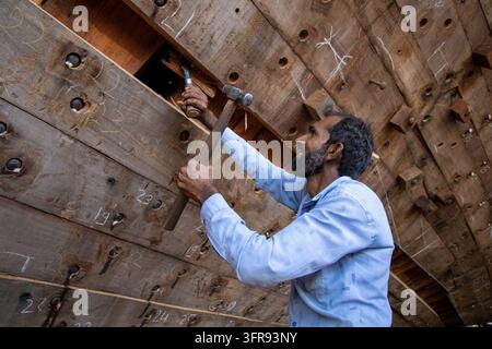 A bearded carpenter hammers a wooden peg into a plank marked with chalk numbers inside a dhow hull, Gujarat, Mandvi, India Stock Photo