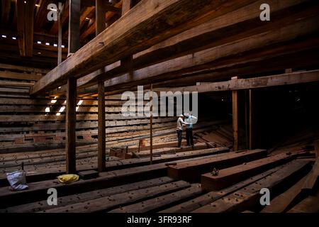 Two shipwrights stand together inspecting the wooden ribs inside the hull of a large ship under construction in a shipyard, Gujarat, Mandvi, India Stock Photo