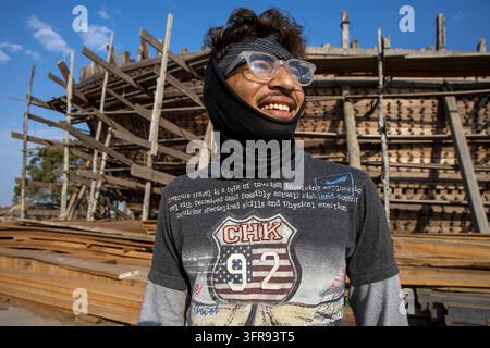 A smiling shipyard worker wearing glasses and a face covering poses in front of a wooden ship under construction, Gujarat, Mandvi, India Stock Photo
