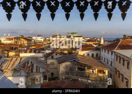 Old town rooftops seen through a carved black wooden window screen at golden hour with rusty corrugated iron roofs and the sea on the horizon, Zanziba Stock Photo