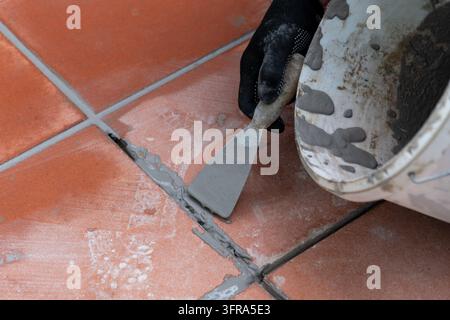 A gloved hand uses a putty knife to apply gray grout between terracotta floor tiles. Stock Photo