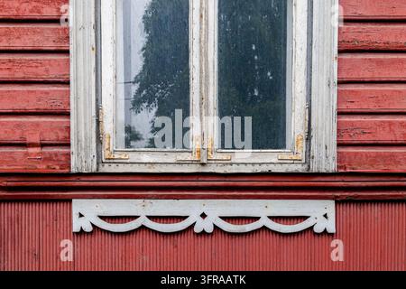 Weathered white wooden window frame with decorative fretwork trim on a red timber clapboard wall, with a tree reflected in the glass, Estonia Stock Photo