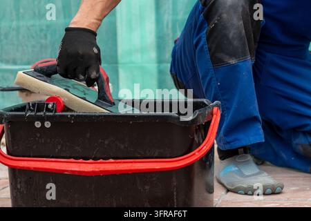 A worker in blue overalls cleans a grout float inside a black bucket. Stock Photo