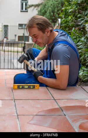 A worker in blue overalls uses a mallet and level while repairing outdoor patio tiles. Stock Photo