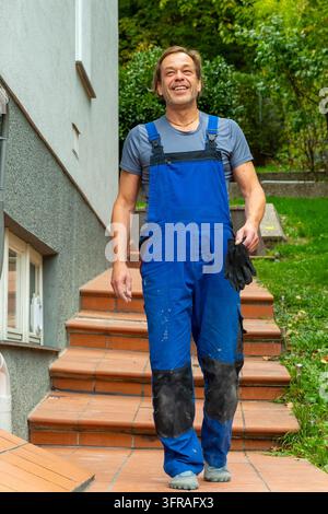 A smiling handyman walks down outdoor steps wearing blue overalls and holding black gloves. Stock Photo
