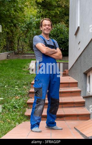 A smiling handyman in blue overalls stands on outdoor steps with arms crossed. Stock Photo