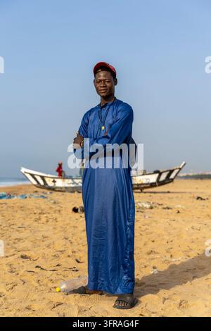 Portrait of a young man wearing a traditional blue robe standing on a beach with fishing boats in the background, Maritime Region, Lome, Togo Stock Photo