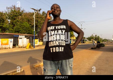 A man wearing a printed tank top laughs while talking on his phone in the middle of a quiet street, Plateaux Region, Kpalime, Togo Stock Photo