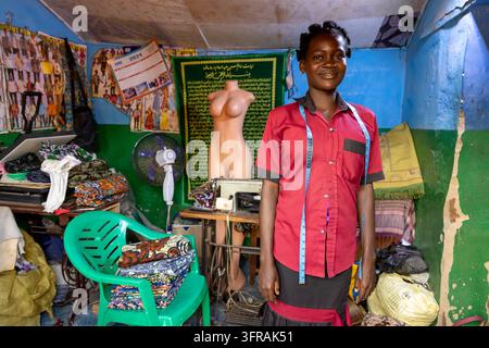 A young seamstress stands in her workshop with a measuring tape around her neck beside a mannequin and sewing machine, Kara Region, Kabye mount, Togo Stock Photo