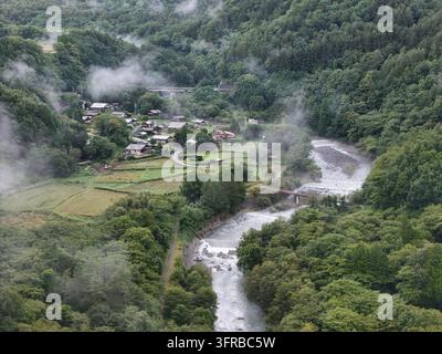 Scattered houses and fields sit beside a curving rocky river as clouds drift across the wooded slopes. Stock Photo
