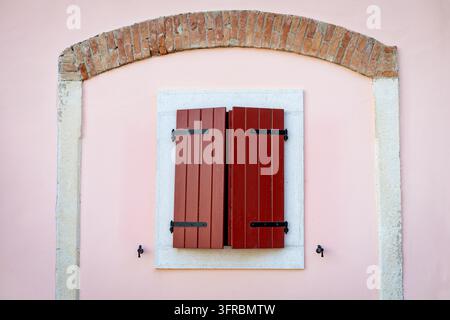 Traditional red wooden window shutters on a pastel pink facade framed by an old rustic brick arch, charming Mediterranean architecture with Stock Photo