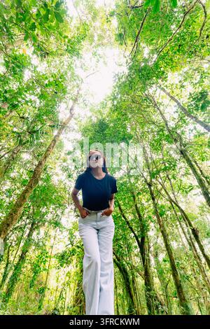 Low angle portrait of a young Asian woman wearing sunglasses standing in a dense rainforest under a tall green tree canopy. Stock Photo