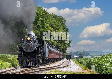 A steam locomotive caresses clouds of smoke as it glides on tracks in a vibrant green landscape The sun shines brightly and fluffy clouds murky the bl Stock Photo