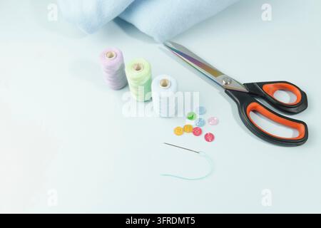 Sewing tools with colorful spools of thread, scissors, buttons, and needle, on a light blue fabric background Stock Photo