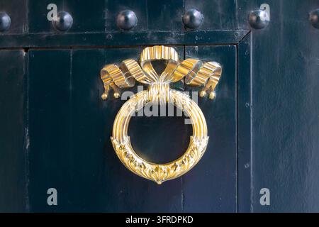 Close-up view of a decorative golden brass ring door knocker with a ribbon bow motif mounted on a dark wooden entrance door. Stock Photo