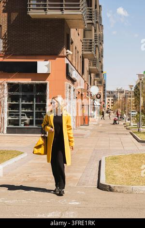 Elegant modern Woman with blond hair in a bright yellow coat and sunglasses walks along a city street. Urban style of modern woman walking shopping Stock Photo