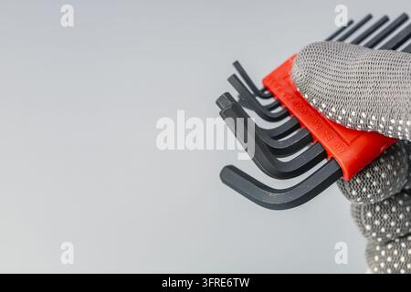 Mechanic hand in gray protective glove holding black hex allen key wrench set in red plastic holder, close-up on gray background with copy space. Stock Photo