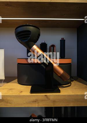 Black and copper hair dryer on wooden shelf. Dryer stands upright on black stand. Background shows other hair styling tools. Shelf wood grain texture Stock Photo