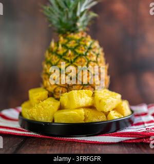 Fresh pineapple chunks served on a black plate with a whole ripe tropical pineapple softly blurred in the background. Stock Photo