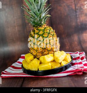 Fresh pineapple chunks served on a black plate with a whole ripe tropical pineapple softly blurred in the background. Stock Photo