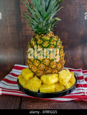 Fresh pineapple chunks served on a black plate with a whole ripe tropical pineapple softly blurred in the background. Stock Photo