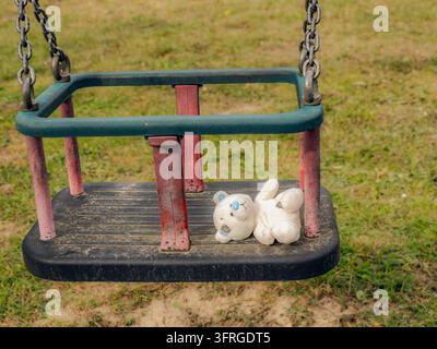 An old white teddy bear lies on a dusty toddler swing at an empty playground, evoking nostalgia, childhood memory and loneliness with worn chains, fad Stock Photo