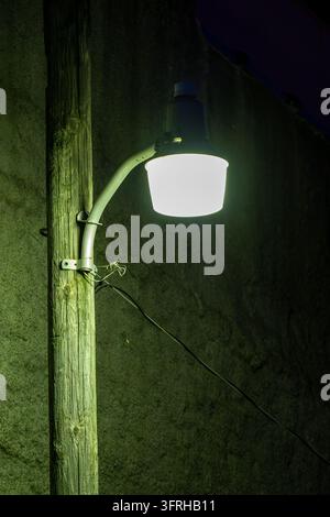 An illuminated security light extends from a curved metal arm on a weathered wooden pole, with an electrical cable crossing the dark background. Stock Photo