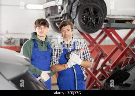 Seasoned middle aged auto repair service owner proudly posing with young adult son, aspiring mechanic continuing his fathers legacy, in workshop fille Stock Photo