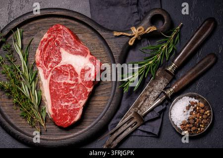 A premium raw T-bone steak is arranged with rosemary, thyme, salt and pepper on a dark rustic surface. Stock Photo