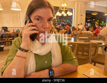 A woman in a green shirt and white scarf speaks on the phone in a warmly lit restaurant with a lively atmosphere in Madrid, Spain. Stock Photo