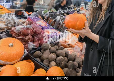 Woman holding an orange pumpkin in a supermarket produce section surrounded by pumpkins, beets, carrots and other fresh vegetables. Seasonal grocery s Stock Photo