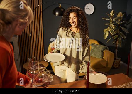 Happy Biracial Woman Setting Dinner Table with Friend at Home Stock Photo