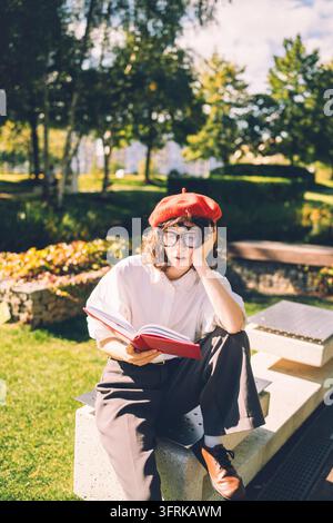 Young student in red beret looks bored reading a book in a park Stock Photo