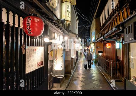 Woman with a rolling suitcase walks a rain-wet lane lined with traditional wooden restaurants in the Pontocho dining district, Kyoto, Japan Stock Photo