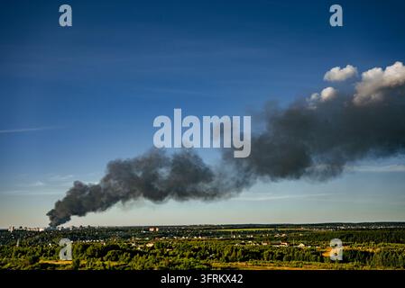 Thick black smoke billows into the clear blue sky, originating from a large fire burning within a residential or industrial area, creating an urban disaster and polluting the environment Stock Photo